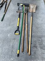 Four used garden tools laid on concrete including a small push broom head, post-hole digger, transfer shovel, and planting shovel, showing wear and rust.