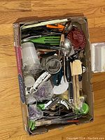 Box filled with assorted kitchen utensils including metal and plastic spatulas, cooking spoons, peelers, bottle openers, stainless steel skewers, plastic measuring cups, and a variety of cookie cutters