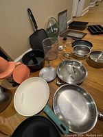 Close-up side view of cookware items showing frying pans, saucepans, glass container, colander, and other kitchen utensils.