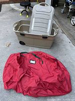 Beige wheeled storage container with lid and large red bag placed on ground outdoors, showing both items fully to depict size and shape.