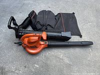 Orange and black Black & Decker leaf blower and mulcher with attached vacuum hose and black collection bag with straps, laid on ground.