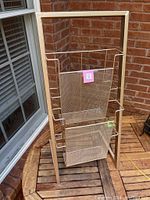 Front perspective showing the entire metal magazine rack with two mesh baskets and wooden frame on a wooden surface near a glass window and brick wall background.
