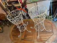 Two white wire planter holders with curved ribbon-like scroll details set on wooden table.