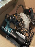 Box containing Makita reciprocating saw, Skilsaw circular saw, and Ryobi drill with cords tangled.