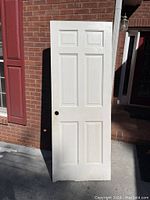 Full view of white solid wood door leaning against wall, showing six panels and round hole for doorknob.