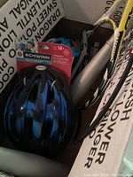 Two bicycle helmets and a bike pump packed together in a box. One helmet is new in package Schwinn brand with blue and black color. The other helmet is used, silver and blue. The bike pump is silver with black hose, Bell brand.