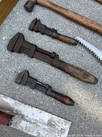 Close-up image showing three vintage monkey wrenches with wooden handles on concrete surface with other tools partially visible.