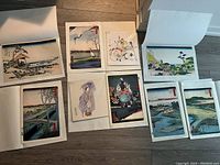 Overview of nine artworks arranged on floor showing Japanese-style prints and other painted/drawn pieces