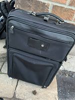 Front view of black Danier leather carry on suitcase showing multiple zippered compartments, handle, and wheels.