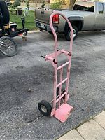 Side view of a pink metal dolly/hand truck showing frame, rounded handle, and deflated wheel