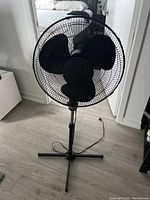 Photo showing black pedestal fan with protective grille, three blades, and cross-shaped base placed on light wood floor.