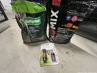 Three garden-related items arranged on a concrete floor: one Golfgreen Tree & Hedge Fertilizer bag, one Promix Premium Organic Vegetable and Herb Mix bag, and one Gardena Quick Connector Starter Kit in package.