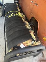 Futon folded with cushions secured by yellow straps, showing black side and patterned upholstery, metal frame visible