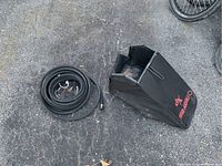 Photo showing a black fabric lawnmower grass catcher bag and a coiled black soaker hose on pavement.