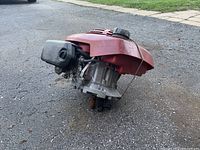 Photo showing side view of the red lawnmower engine with attached black fuel tank and air filter housing on cracked asphalt surface.