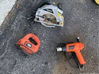 Mastercraft circular saw with metal blade guard, Black & Decker orange jigsaw, and orange certified heat tool on ground asphalt.