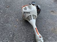 Base motor unit of Stihl Kombi with white and orange housing, pull starter, shaft and handle grip lying on asphalt.