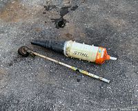 Pair of Stihl Kombi system attachments laid on ground, showing the straight trimmer attachment with metal shaft and blower attachment with plastic housing.