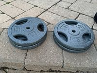 Four 25 lb weight plates shown stacked and side-by-side on outdoor stone surface, highlighting textured dark gray finish and grip holes.