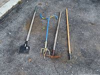Four yard tools laid out on pavement including an edger, manual tiller, and two hoes with wooden handles and metal tool heads.