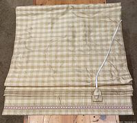 Roman blind folded flat with visible bottom edge beaded trim, braided pull cord with silk tassel on a wooden surface.