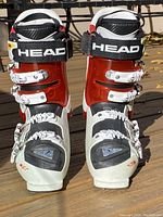 Front view of pair of Head Vector 120 ski boots showing red and white plastic shell, white buckles, black strap with HEAD branding on wooden surface.