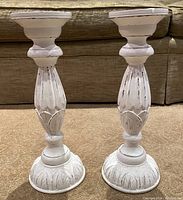 Front view of two large wooden candlesticks showing carved details and distressed white paint finish.