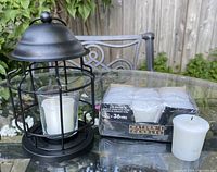 Single black metal outdoor garden lantern with glass candle holder containing a white candle, next to a package of seven unused white candles by Watson's Candles.