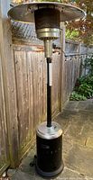 Full view of the propane patio heater showing the stainless steel top reflector, mesh burner, black pole and base with wheels. Positioned outdoors next to a wooden fence.