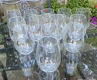 Thirteen clear plastic stemless wine glasses grouped together on an outdoor table, each with gold 'Cheers' lettering on front.