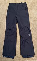 Front view of black Rossignol boys ski pants laid flat on carpet. Shows full legs, zippered pockets, and general condition.
