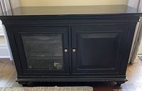 Front view of black TV console showing one glass door and one solid door with metal knobs, and visible shelves inside the glass door.