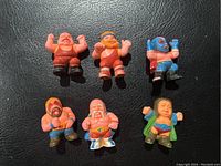 Front view showing all 6 vintage rubber wrestler figures lying on black textured surface, displaying various colors and poses