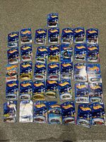 Overview photo showing 36 Hot Wheels cars in original blister card packaging laid out on carpet. Cards have blue backgrounds with Hot Wheels logo.
