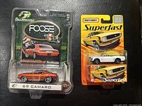 Photo shows two diecast Camaro models still in original packaging: orange Foose 69 Camaro and white Matchbox Camaro SS Superfast series.