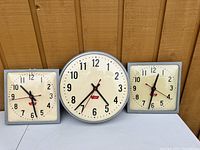 Front view of three old high school clocks, one large round clock between two smaller square clocks with curved glass and metal frames.