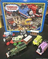 Photo of multiple Thomas & Friends toy vehicles displayed in front of a colorful blue Thomas & Friends tin box with lid showing character artwork.