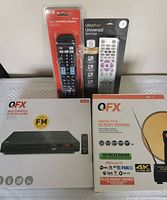 Set of all items on table: QFX multimedia DVD player box with FM radio, two universal remote controls in original packaging (QFX 8-in-1 and UltraPro 6-device), QFX digital TV and HD ready antenna box.