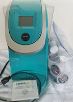 Front view of Keurig 2.0 coffee machine showing teal body, digital display and gathered K-Cup pods in mesh bag alongside instruction manual on white cloth
