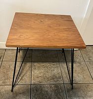 Photo of a square accent table with a natural wood grain top and four black metal hairpin legs, placed on tile flooring against a white wall.