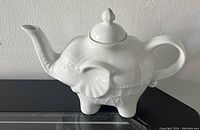 Side view of white ceramic teapot shaped like an elephant with detailed ears and trunk forming the spout, handle, and lid with finial.