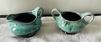 Photo showing a green ceramic cream pitcher with one handle and a green ceramic sugar bowl with two handles, both with a glazed finish, placed on a white cloth against a wall.