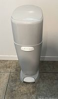 Front view of white Diaper Genie diaper disposal system with foot pedal and lid closed, placed on tiled floor against a white wall.