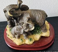 Photo shows a figurine with one adult elephant and two smaller elephants positioned on a textured base, on a reddish wooden platform, viewed from above and front side.