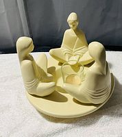 Front view of cream-colored ceramic candle holder featuring a circle of three abstract seated human figures with smooth minimalist design, surrounding a glass votive holder at center.