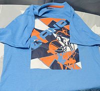Front view of the light blue AND1 T-shirt with bold multi-colored graphic design including 'AND1' text and athlete figure.