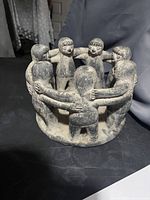 Stone sculpture showing seven figures in a circle with arms interlocked, viewed from an angle.