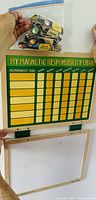 View of the green and yellow magnetic responsibility chart with a hand holding a sealed plastic bag containing assorted wooden magnetic chore pieces above it, and a blank whiteboard framed in wood below the chart.