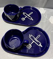 Two sets of deep blue ceramic plates each with an integrated cup holder and matching cups. Plates have white designs of a fork and rolling pin crossed.