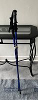 Pair of blue walking sticks leaning against a black table, showing handles, wrist straps, and rubber tips on a carpeted floor.